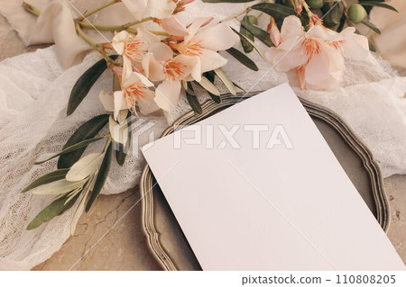 Closeup of blank greeting card, invitation mockup on vintage silver plate. Summer wedding, birthday mock-up. Oleander blossoms, muslin textile. Olive tree branches. Blurred marble background. Top. Closeup of blank greeting card, invitation mockup on vintage silver plate. Summer wedding, birthday mock-up. Oleander blossoms, muslin textile. Olive tree branches. Blurred marble background. Top. 110808205