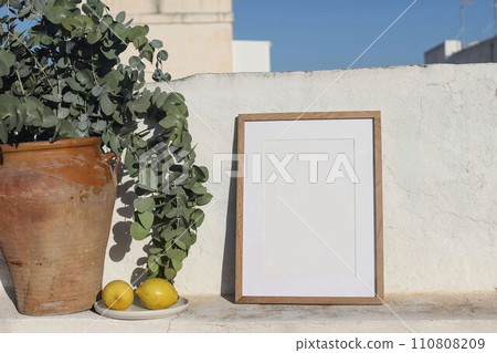Blank vertical wooden frame picture mock up. Vintage ceramic vase with eucalyptus branches, fresh lemons fruit. White textured white wall in sunlight. Summer holiday display background. Art, poster. 110808209