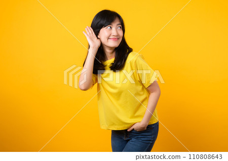 Capture attention portrait of young Asian woman. Wearing a yellow t-shirt and denim jeans, leans in to overhear and listen, evoking curiosity and intrigue. attention-grabbing promotions and discounts. Capture attention portrait of young Asian woman. Wearing a yellow t-shirt and denim jeans, leans in to overhear and listen, evoking curiosity and intrigue. attention-grabbing promotions and discounts. 110808643