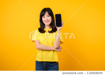 Experience joy with cheerful portrait, young Asian woman wearing yellow t-shirt and denim jeans showcases happy smile while showing smartphone screen. concept of recommended applications. Experience joy with cheerful portrait, young Asian woman wearing yellow t-shirt and denim jeans showcases happy smile while showing smartphone screen. concept of recommended applications. 110808649