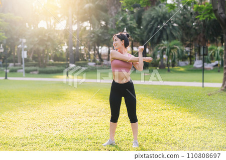 wellness and healthy lifestyle portrait of 30s Asian woman in pink sportswear. preparing and stretching body and arm muscles before sunset run in park. fitness outside and embrace balanced life. wellness and healthy lifestyle portrait of 30s Asian woman in pink sportswear. preparing and stretching body and arm muscles before sunset run in park. fitness outside and embrace balanced life. 110808697