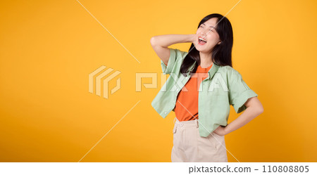 essence of happiness and well-being with cheerful young Asian woman in her 30s wearing an orange shirt. Her happy mind wellness gesture on yellow background illuminates compelling happiness portrait. essence of happiness and well-being with cheerful young Asian woman in her 30s wearing an orange shirt. Her happy mind wellness gesture on yellow background illuminates compelling happiness portrait. 110808805