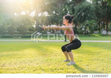 Healthy outdoor lifestyle with fit young Asian woman 30s wearing pink sportswear squat muscles in public park before going for invigorating run, embodying the wellness concept. Healthy outdoor lifestyle with fit young Asian woman 30s wearing pink sportswear squat muscles in public park before going for invigorating run, embodying the wellness concept. 110808807