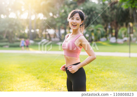 Experience the joy of wellness living as a happy, beautiful 30s Asian woman looks at the camera with a smile in a park at sunset. 110808823