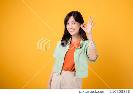 charming young Asian woman 30s, elegantly attired in orange shirt and green jumper. Her captivating okay hand gesture and gentle smile shine against a yellow background, body language concept. 110808828