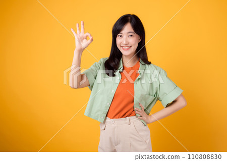 young Asian woman 30s, elegantly clad in orange shirt and green jumper. Her endearing okay hand gesture and gentle smile, set against a yellow background, reveal the beauty of body language. young Asian woman 30s, elegantly clad in orange shirt and green jumper. Her endearing okay hand gesture and gentle smile, set against a yellow background, reveal the beauty of body language. 110808830