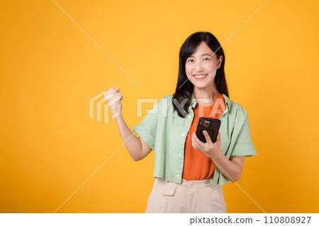 Excited young Asian woman in her 30s, wearing orange shirt and green jumper, using smartphone with fist up gesture on yellow studio background. Great news from mobile concept. 110808927