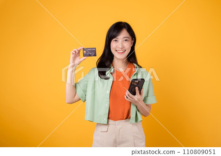 young Asian woman in her 30s, dressed in orange shirt and green jumper, shops online using smartphone with credit card on yellow studio background. Mobile concept. young Asian woman in her 30s, dressed in orange shirt and green jumper, shops online using smartphone with credit card on yellow studio background. Mobile concept. 110809051
