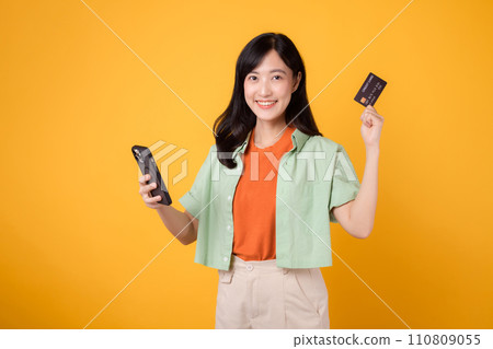 convenience of shopping from mobile with young Asian woman in her 30s, elegantly dressed in orange shirt and green jumper, using smartphone while showcasing credit card on yellow studio background. convenience of shopping from mobile with young Asian woman in her 30s, elegantly dressed in orange shirt and green jumper, using smartphone while showcasing credit card on yellow studio background. 110809055