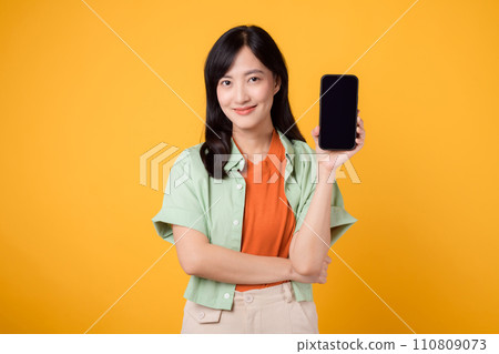 Happy young Asian woman 30s, wearing orange shirt and green jumper, displays smartphone screen on yellow studio background. New mobile application, online shopping, and new mobile device concept. Happy young Asian woman 30s, wearing orange shirt and green jumper, displays smartphone screen on yellow studio background. New mobile application, online shopping, and new mobile device concept. 110809073