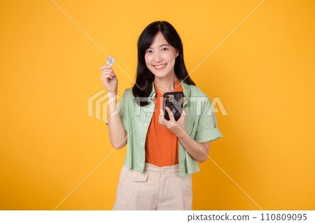 future of finance with cheerful young Asian woman in her 30s, donning orange shirt and green jumper, displaying smartphone screen and crypto currency coin on yellow background. Future finance concept. future of finance with cheerful young Asian woman in her 30s, donning orange shirt and green jumper, displaying smartphone screen and crypto currency coin on yellow background. Future finance concept. 110809095