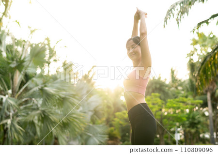 fit 30s young Asian woman in pink sportswear plays yoga and stretches muscles. Embrace healthy outdoor lifestyle with fitness yoga girl in public park at sunset. wellness and well being concept. 110809096