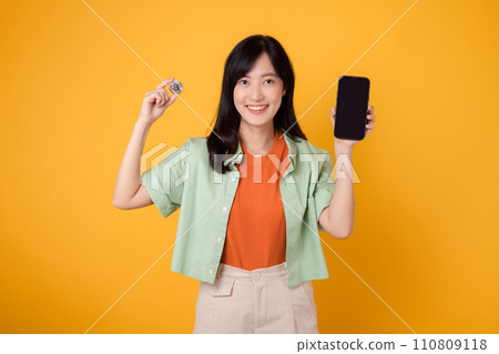 the future of finance with a captivating young Asian woman in her 30s, elegantly dressed in orange shirt and green jumper, using smartphone screen and crypto currency coin on yellow background. 110809118
