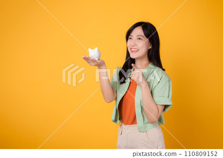 Happy young Asian woman in her 30s, wearing orange shirt and green jumper, showcases piggy bank while pointing finger to free copy space on yellow background. Financial money concept. 110809152