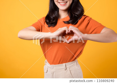 healthcare, wellness, and well-being with young Asian woman 30s wearing an orange shirt. Witness heart hand gesture on her chest against a vibrant yellow background, concept of holistic well-being. healthcare, wellness, and well-being with young Asian woman 30s wearing an orange shirt. Witness heart hand gesture on her chest against a vibrant yellow background, concept of holistic well-being. 110809240