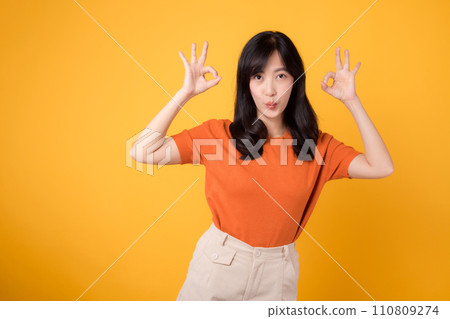 In a vibrant yellow backdrop, a young Asian woman in her 30s, donning an orange shirt, displays the okay sign gesture. Hands gesture concept. In a vibrant yellow backdrop, a young Asian woman in her 30s, donning an orange shirt, displays the okay sign gesture. Hands gesture concept. 110809274