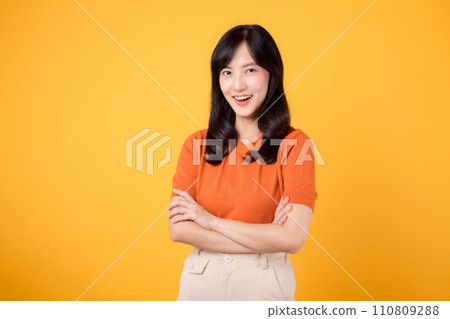 Step into confident grace with a young Asian woman in her 30s, wearing an orange shirt, displaying crossed arm sign gesture on yellow background. Step into confident grace with a young Asian woman in her 30s, wearing an orange shirt, displaying crossed arm sign gesture on yellow background. 110809288