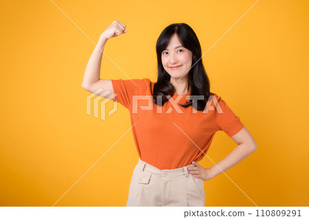 Embrace confidence and feminist empowerment as a young Asian woman in her 30s raises a fist up hand sign gesture, wearing an orange shirt on yellow background. Empowerment and feminism concept. Embrace confidence and feminist empowerment as a young Asian woman in her 30s raises a fist up hand sign gesture, wearing an orange shirt on yellow background. Empowerment and feminism concept. 110809291