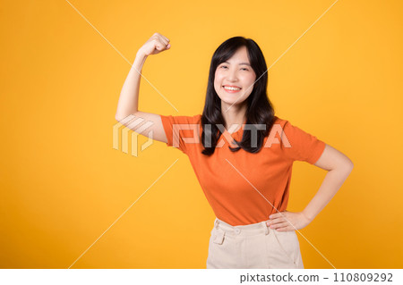 Feel empowered with a young Asian woman in her 30s celebrating a fist up hand sign gesture, wearing an orange shirt on yellow background. Empowerment and feminism concept. Feel empowered with a young Asian woman in her 30s celebrating a fist up hand sign gesture, wearing an orange shirt on yellow background. Empowerment and feminism concept. 110809292