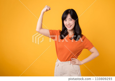 Confident young Asian woman in her 30s raises a fist up hand sign gesture, wearing an orange shirt on yellow background. Empowerment and celebration concept. Empowerment and feminism concept. Confident young Asian woman in her 30s raises a fist up hand sign gesture, wearing an orange shirt on yellow background. Empowerment and celebration concept. Empowerment and feminism concept. 110809293