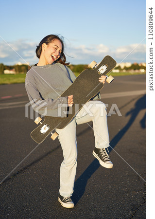 Funny asian girl enjoying skating, holding skateboard like guitar and shadow playing, having fun outdoors Funny asian girl enjoying skating, holding skateboard like guitar and shadow playing, having fun outdoors 110809644