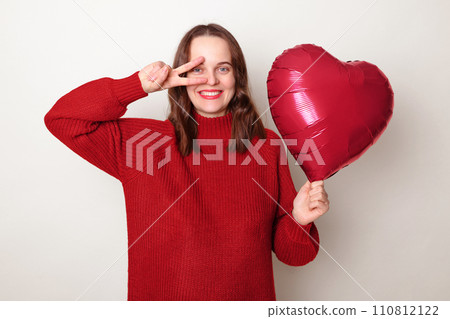 Cheerful smiling brown haired adult woman wearing red sweater holding heart shape balloon posing isolated over gray background enjoying festive event showing v sign near eyes rejoicing 110812122