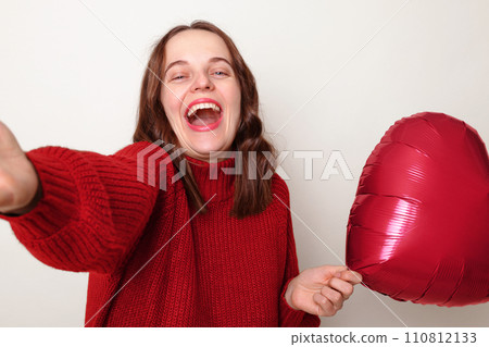 Extremely happy Caucasian woman wearing red sweater holding heart shaped balloon isolated over gray background making point of vie photo laughing happily 110812133