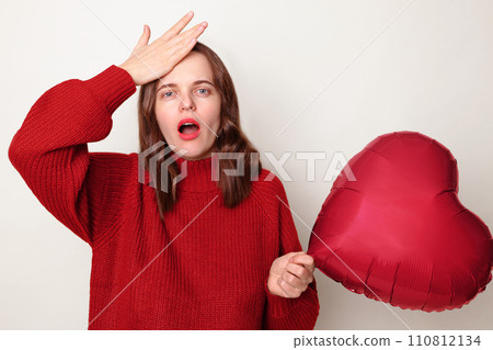 Shocked amazed woman with brown hair wearing red jumper standing isolated over gray background with heart shaped balloon showing facepalm gesture feeling sorrow regret blaming herself for failure 110812134