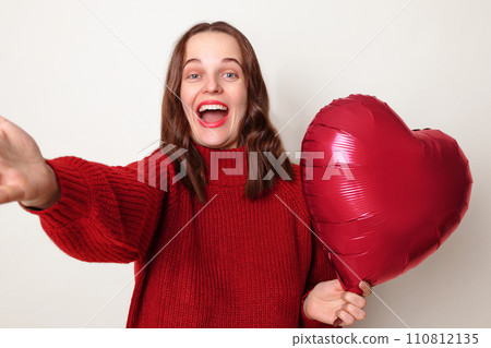 Overjoyed woman with brown hair wearing red jumper standing isolated over gray background with heart shaped balloon making pov selfie having excite amazed expression 110812135
