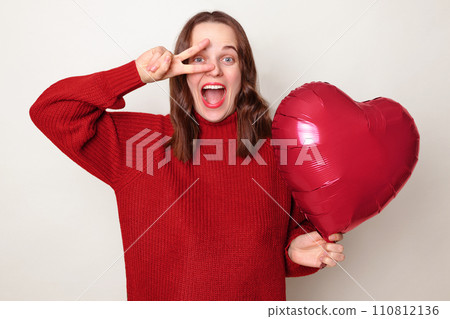 Excited festive woman with brown hair wearing red jumper making victory sign in front her eyes having fun standing isolated over gray background with heart shaped balloon 110812136