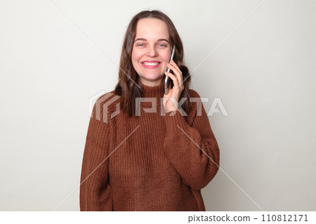 Cheerful Caucasian woman wearing brown jumper talking on mobile phone enjoying pleasant conversation looking smiling at camera standing isolated over gray background 110812171