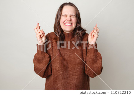 Attractive brown haired adult woman wearing brown sweater posing isolated over gray background standing with crossed fingers and closed eyes praying for good luck making wish 110812210