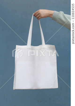 Blank white tote bag canvas fabric with handle mock up design. Close up of woman hand holding eco or reusable shopping bag on blue metal wall. No plastic bag and ecology concept. Vertical. 110814535