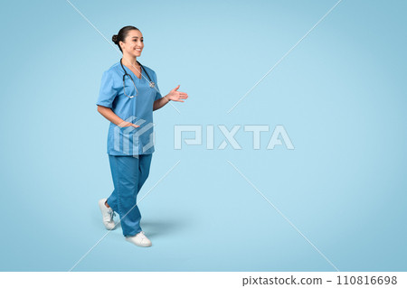 Cheerful young female nurse in blue scrubs with stethoscope around her neck, walking and stretching hand for greeting 110816698