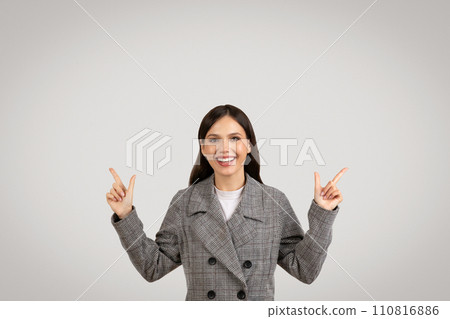 Cheerful woman in plaid jacket pointing upwards with both hands 110816886