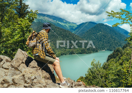 Hiking traveller caucasian bearded man with backpack sitting on rocks enjoying beautiful view of turquois blue Lake Ritsa in Abkhazia 110817764