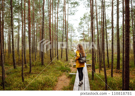 Beautiful woman in yellow raincoat walks along wooden path in forest. Concept of recreation, tourism 110818781