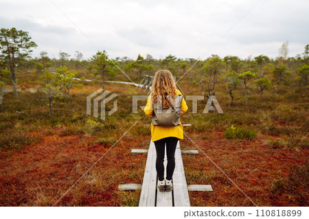 Woman traveler in a yellow coat walks along a wooden walking path along the wetlands. Back view. Woman traveler in a yellow coat walks along a wooden walking path along the wetlands. Back view. 110818899