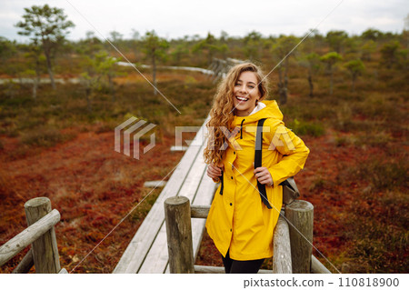Smiling female traveler in yellow coat walks along wooden path among wild nature. Travel, vacation. Smiling female traveler in yellow coat walks along wooden path among wild nature. Travel, vacation. 110818900