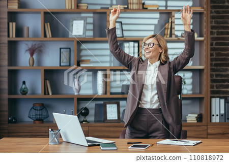 Joyful mature professional woman with arms raised in triumph at a stylish home office setup, exuding confidence and success. 110818972