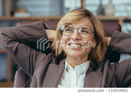 Successful blonde senior businesswoman with glasses relaxing and smiling in her stylish home office, embodying professionalism and confidence. 110818981