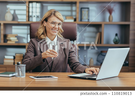 Smiling mature businesswoman with glasses working on laptop in home office, holding a glass of water reflecting a healthy lifestyle. 110819013