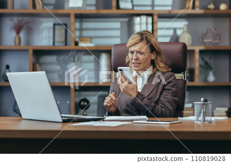 A senior businesswoman appears concerned while multitasking at her home office, working on laptop and using smartphone. 110819028