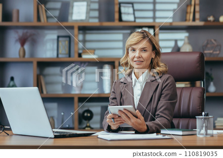 Professional mature businesswoman in a stylish home office setup, using a tablet with focused expression. 110819035