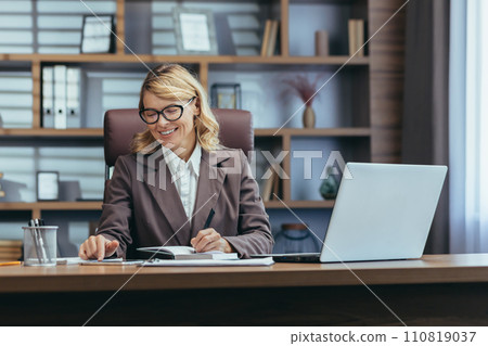 Elegant senior woman in business attire smiles while working at her laptop in a well-appointed home office setup, exuding confidence. 110819037