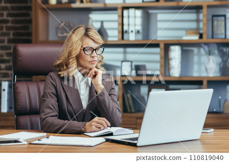 Elegant senior businesswoman concentrating while working at her home office with a laptop, pen, and notebook. Elegant senior businesswoman concentrating while working at her home office with a laptop, pen, and notebook. 110819040