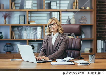 A professional senior businesswoman with blonde hair focused on work at her laptop in a well-organized home office. A professional senior businesswoman with blonde hair focused on work at her laptop in a well-organized home office. 110819049