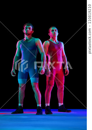 Two young confident and focused men in blue and red sportwear posing against black background in mixed neon lights. Two young confident and focused men in blue and red sportwear posing against black background in mixed neon lights. 110819210