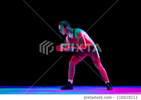 Freestyle wrestler in red fighting tights stands in attack position and looking ta camera against black background in mixed neon lights. 110819211
