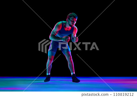 Sportsman, freestyle wrestler in blue fighting tights stands in attack position and looking ta camera against black background in mixed neon lights. Sportsman, freestyle wrestler in blue fighting tights stands in attack position and looking ta camera against black background in mixed neon lights. 110819212